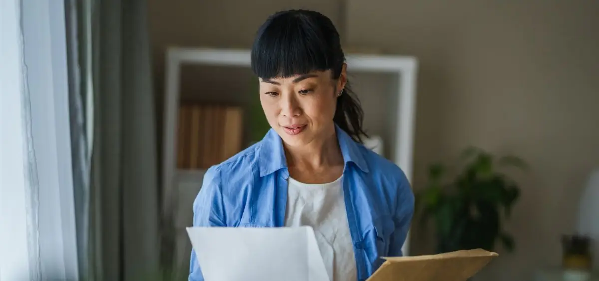 A woman is standing in her living room holding a large envelope and reading the papers that were inside.