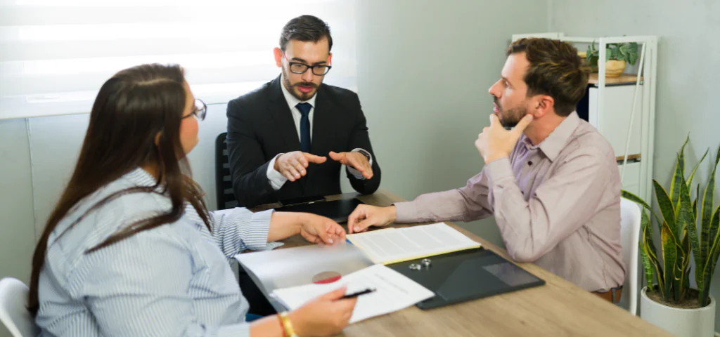 Trois personnes sont assises autour d’une table dans un bureau clair. Une personne portant un veston semble expliquer quelque chose avec ses mains. Les deux autres personnes semblent l’écouter.