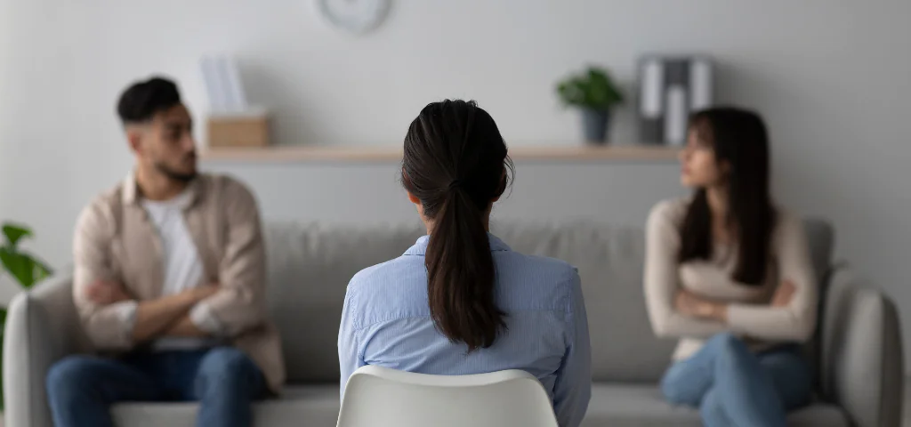 Une femme et un homme sont assis chacun à l’extrémité d’un canapé. De dos en avant-plan, une médiatrice se trouve visuellement entre les deux, cherchant à trouver un terrain d’entente.