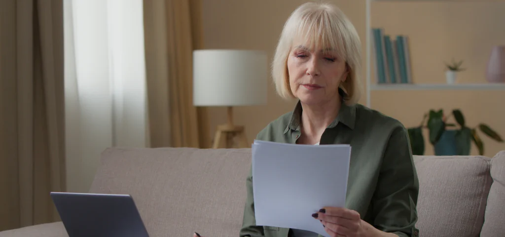 Une femme aînée lit des documents assise sur son canapé.