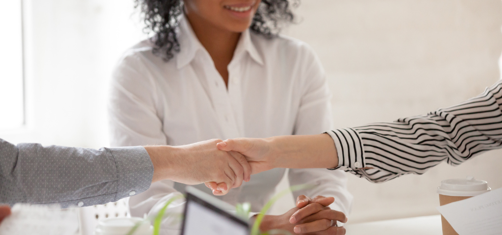 Deux personnes se serrent la main au-dessus d’une table. Une troisième personne, assise avec les mains jointes, les observe en souriant.