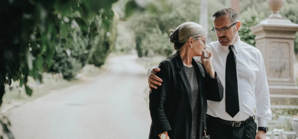 A husband comforts his grieving wife after a funeral.
