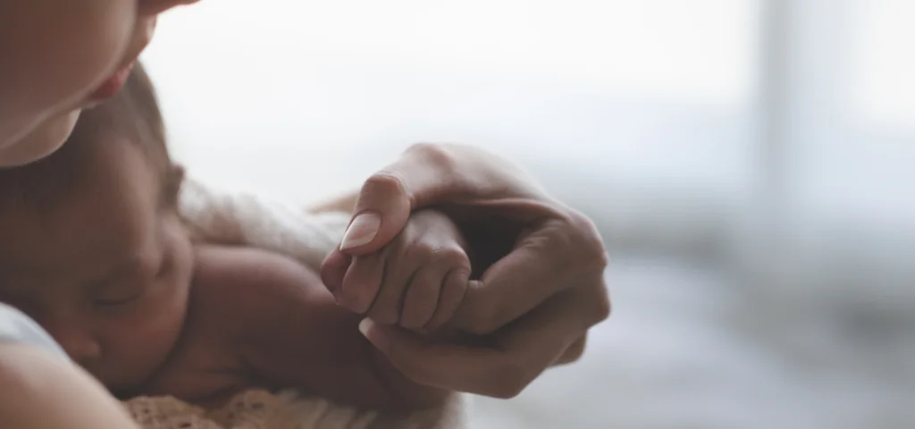 A woman holds a small baby close to her chest. The baby is holding on to one of the woman's fingers.