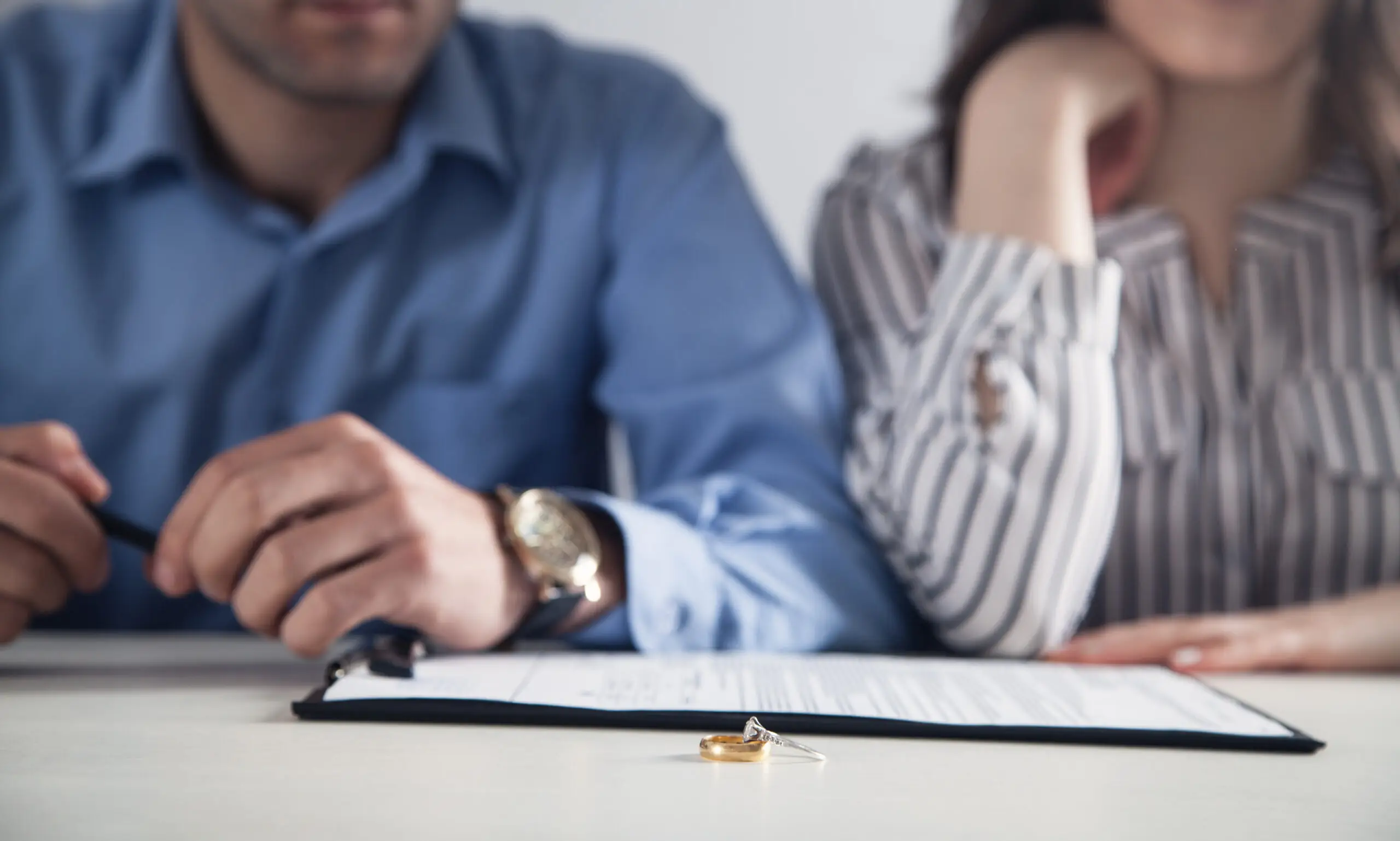 A man and a woman sit side by side at a table with a document in front of them. Two wedding rings rest on the document.