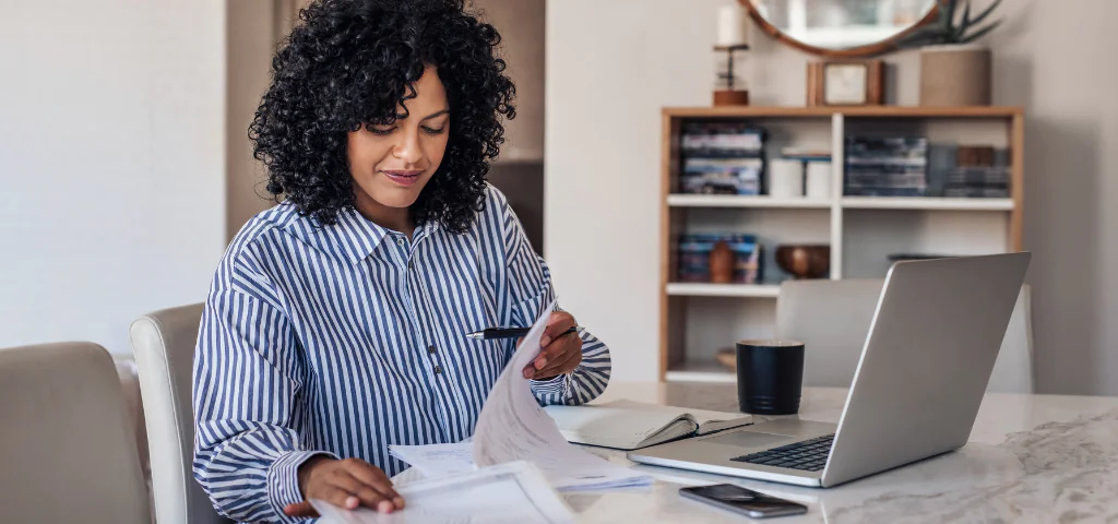 Une femme est assise à une table et lit des documents. Un ordinateur portable est ouvert devant elle.