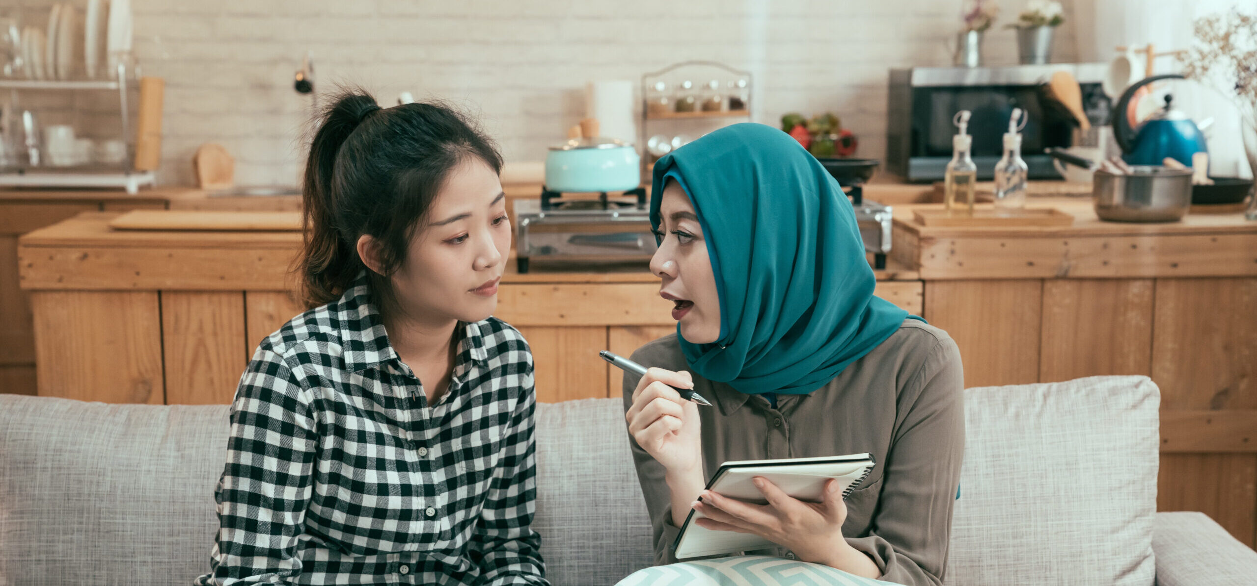Two young women are seated on a couch in a kitchen. One wears a hijab and is holding a notebook and pen, appearing to explain something to the other, who listens attentively.