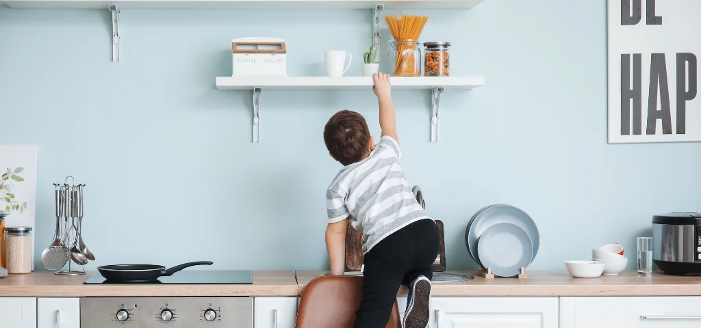 Un enfant est en équilibre sur une chaise et tente d’attraper quelque chose sur une étagère dans la cuisine.