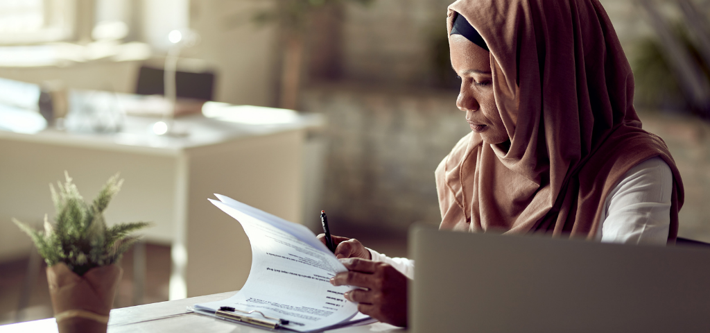 Une femme est assise à une table devant un ordinateur portable. Elle relit un document et tient un stylo dans sa main droite.