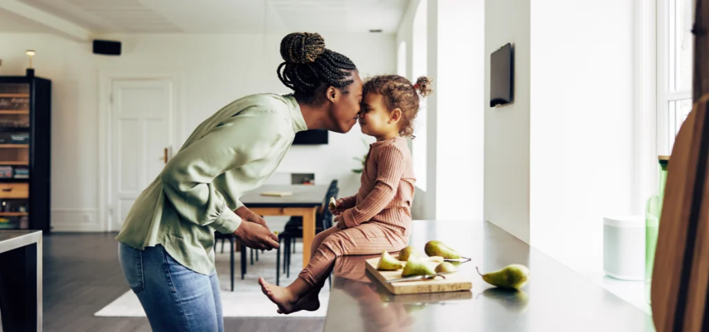 Une personne se penche pour embrasser un enfant assis sur un comptoir de cuisine. Il y a quelques tranches de poires sur le comptoir.

À l’arrière-plan, on voit une table à manger, des chaises et de grandes fenêtres.