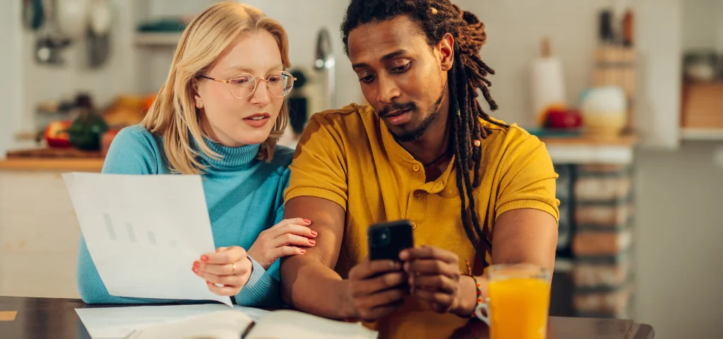 Un couple assis à une table discute de démarches administratives, un téléphone portable à la main et des dossiers posés devant eux.