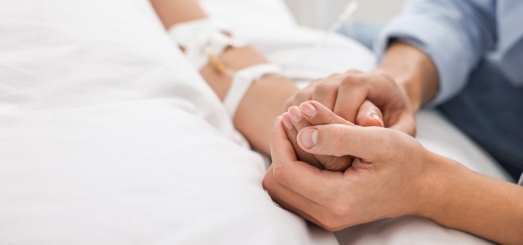 A person gently holds the hand of a patient lying in a hospital bed. The patient has an intravenous catheter attached to their arm, and the scene conveys a moment of support and comfort.