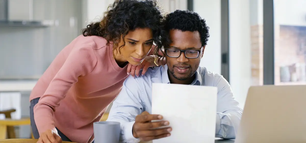 A man sits at a computer while a woman leans over his shoulder. They are both looking at a document.