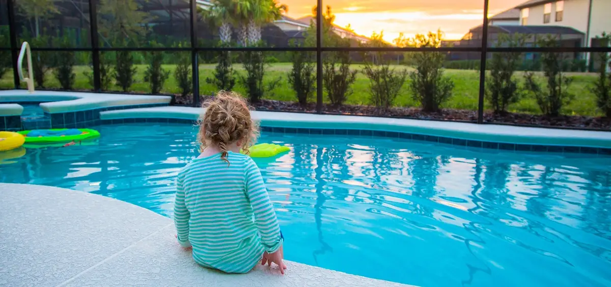 Pool fences must meet new safety standards. Rear view of a child sitting on the edge of a backyard swimming pool.
