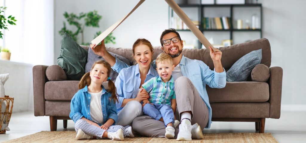 Are you in a parental union? A smiling family sits together on a rug in a bright living room. The parents hold a large piece of cardboard above their heads, forming a symbolic roof over themselves and their two young children.