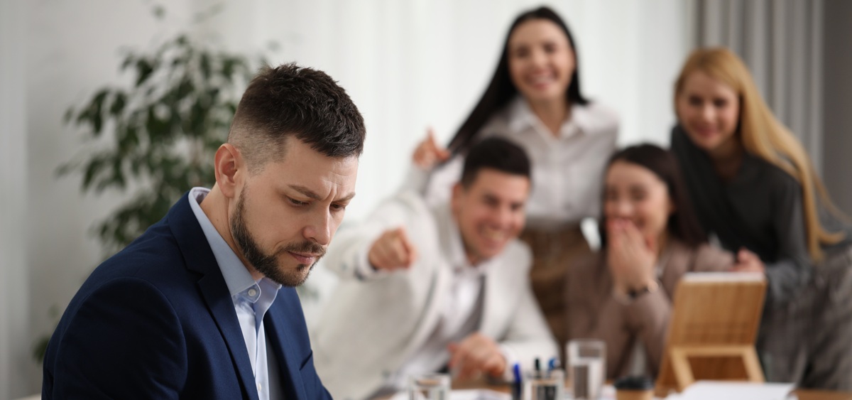 Au premier plan, un homme est assis seul d’un côté d’une table de bureau, l’air préoccupé. En arrière-plan, quatre personnes floues, assises de l’autre côté de la table, rient et le montrent du doigt.