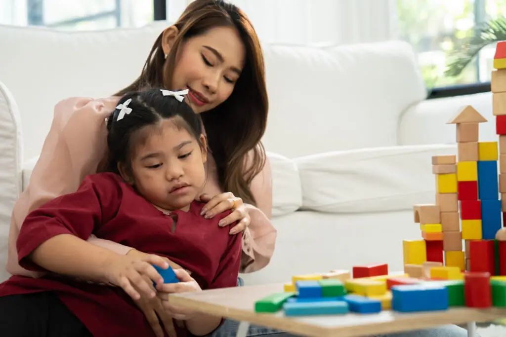 A woman is sitting on a white couch, holding a young girl in her arms. The girl appears to be low-energy or sad.