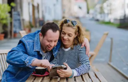 Une femme avec une personne vivant avec le syndrome de l'autisme regardent ensemble un téléphone sur une terrasse près d'un café.