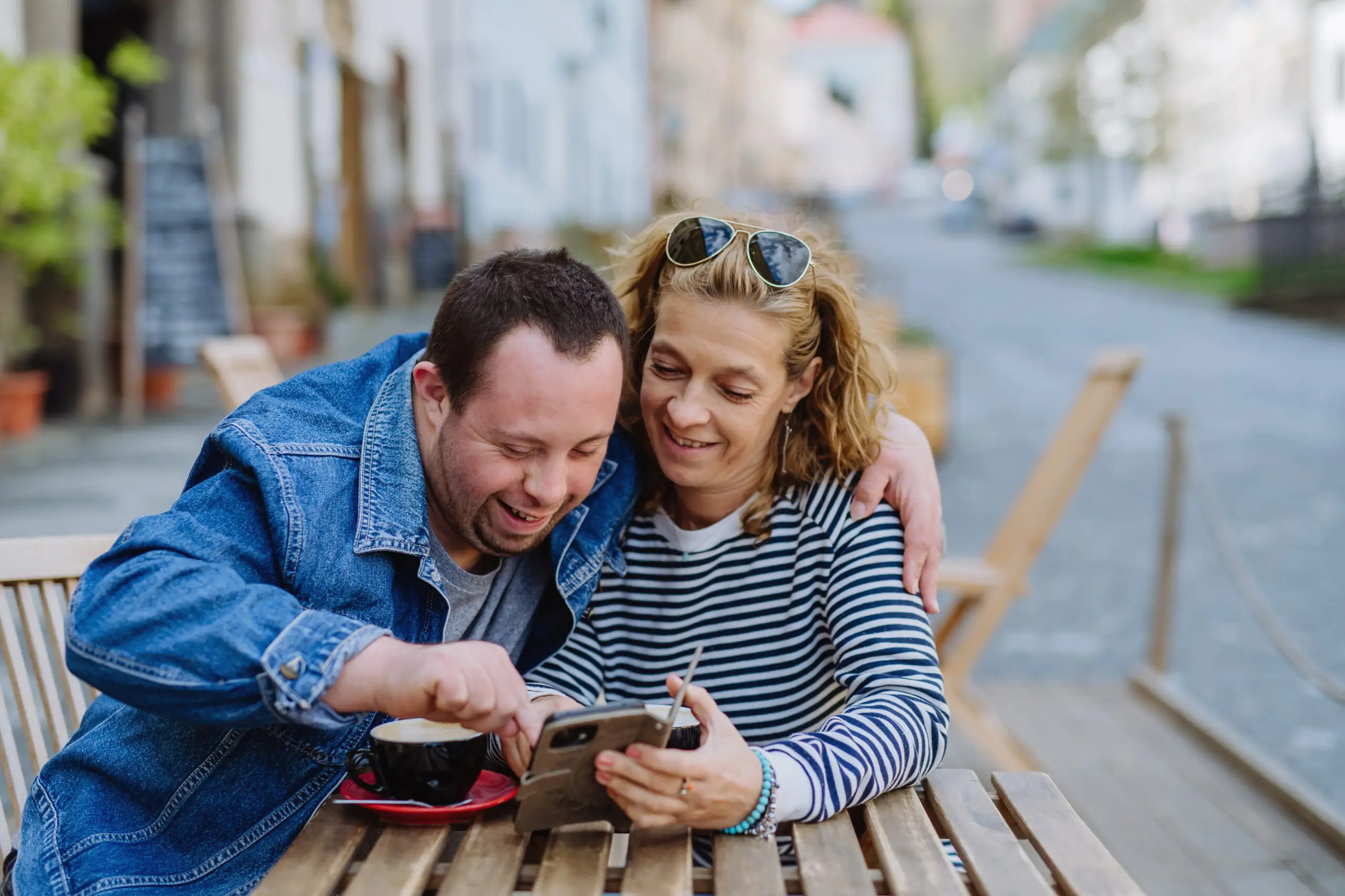 A women with a man living with down syndrome are watching cheerfuly a phone. They are close and enjoy eachother presence.